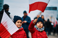 Zdjęcie przedstawia dzieci w czerwonych kurtkach zimowych  i czapkach podczas marszu niepodległości na uliach miasta. Dziewczynki trzymają w ręku flagi biało-czerowne, którymi machają. W tle zarys spacerujących ludzi i budynków
