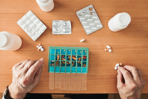 Stół na którym leżą lekarstwa w opakowaniach oraz pudełkach. Widoczne są również dłonie człowieka wyliczające tabletki. 